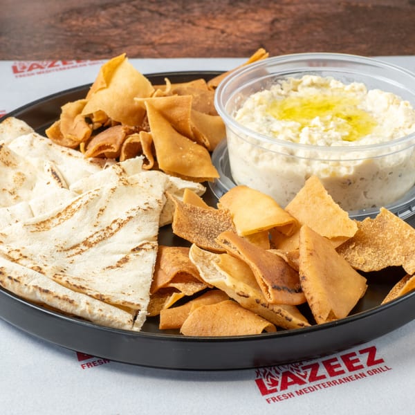 A black plate holds pita bread, fried pita chips, and a plastic container of hummus with olive oil.