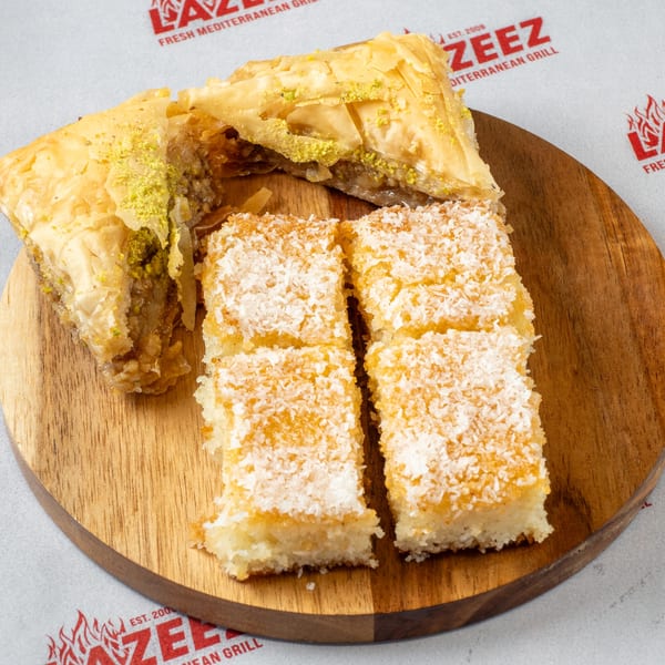 Two pieces of baklava with pistachio and four coconut semolina cakes on a wooden board.