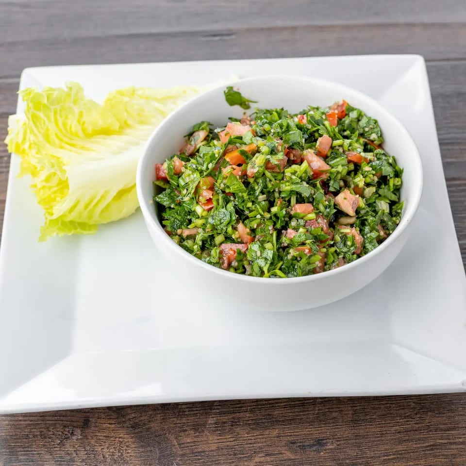 A white bowl of Lazeez Fresh Mediterranean Grill's tabbouleh salad with lettuce on a white plate.